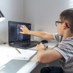 An elementary school student sits at a desk in front of a laptop and communicates with a friend via video link online, at home.