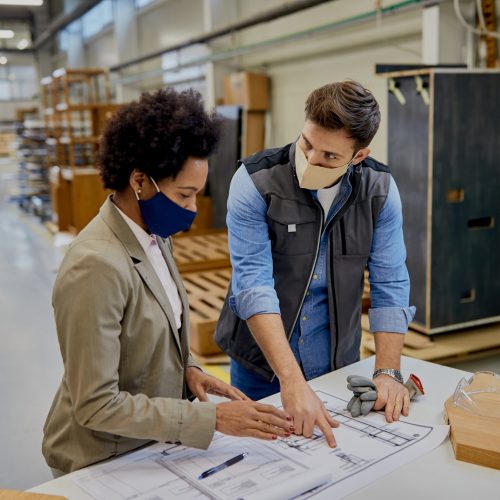 Woodworking foreman and black female company manager examining blueprints in a factory during coronavirus pandemic.